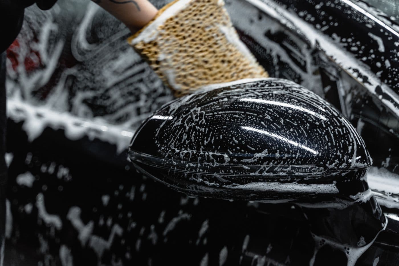 Wash mitt cleaning a black luxury-car mirror covered in soap and water.