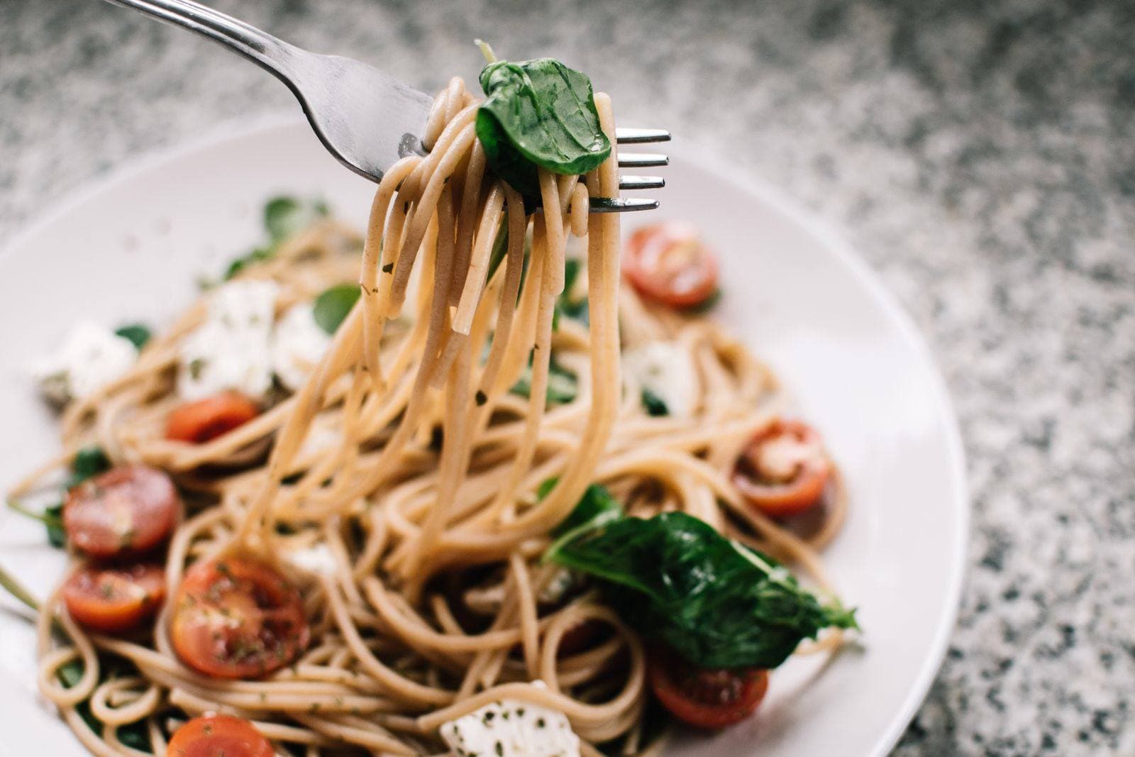 A fork lifting pasta with tomato, basil, and white cheese from a white plate.