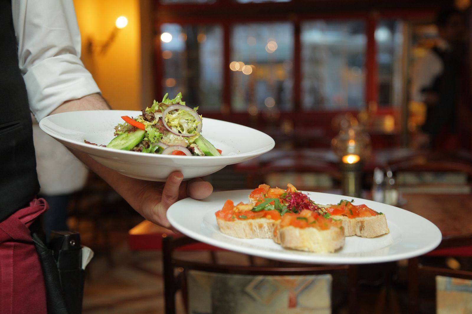 Two plated dishes, a salad and tomato toasts, carried through a dining room.